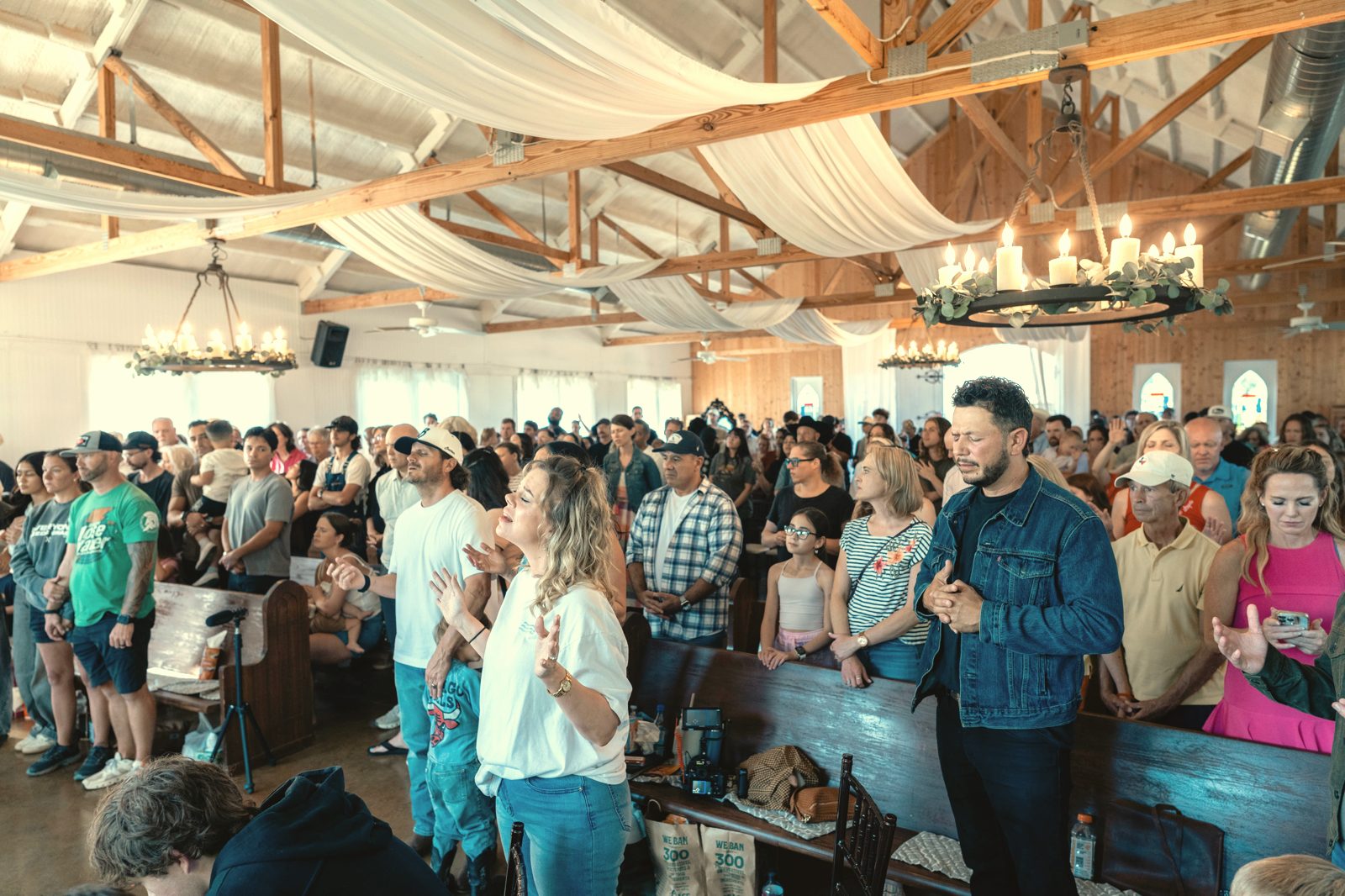 A full barn with wooden rafters and chandeliers as families stand in worship at a Tend Gathering