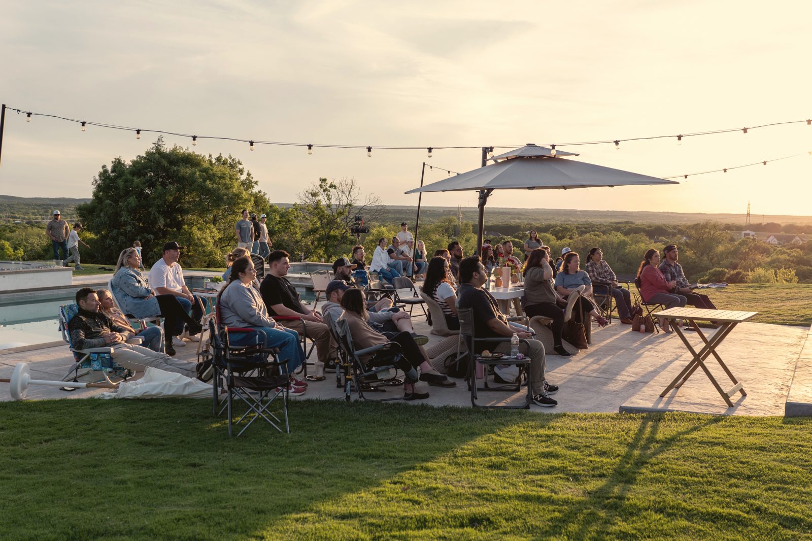 Families seated outdoors under string lights at sunset for a Tend Gathering on open land