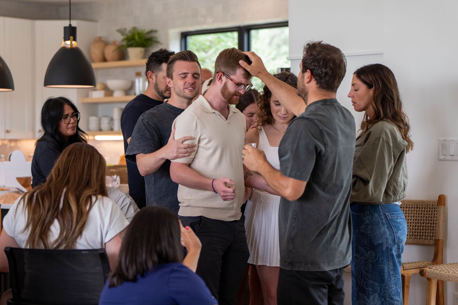 Friends gathered in a bright home kitchen, laying hands on a young man and praying over him