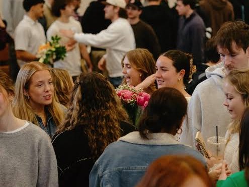 A crowded room of young adults smiling and talking together, some holding flowers