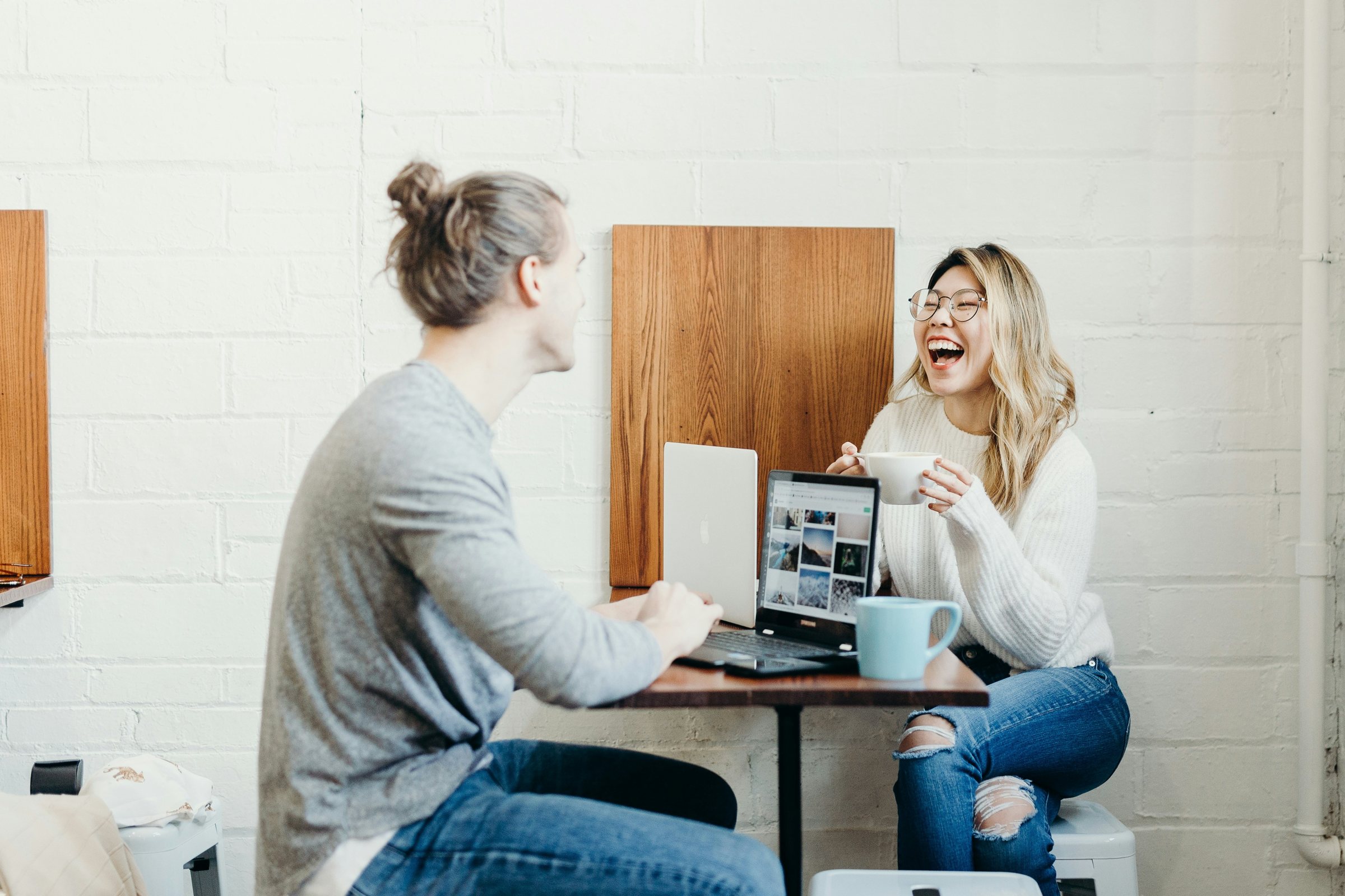 A young man and a young woman laughing together over coffee and laptops in a coffee shop