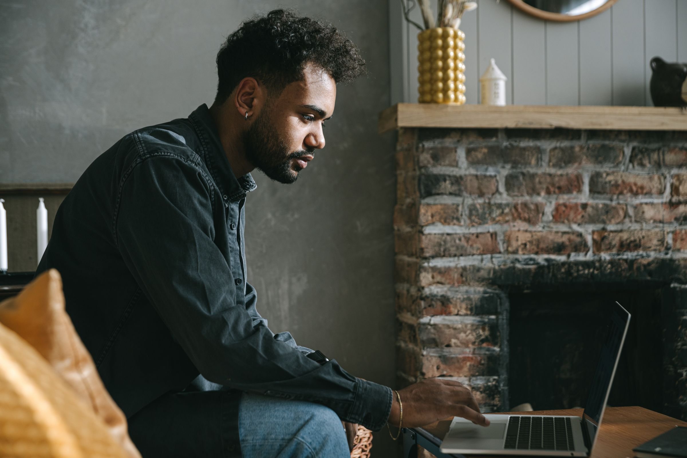 A man alone in his living room by a brick fireplace, joining in on his laptop